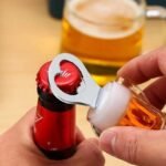 Close-up of hands using a metal bottle opener to pry the red cap off a small orange bottle, with a glass of beer blurred in the background. - Bling On Decor® | Luxury Handmade Home Decor ANJU BALA