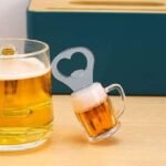 Beer glass with frothy top and a small matching beer mug on a wooden table; a metal bottle opener with a heart-shaped cutout sits nearby, teal box in the background. - Bling On Decor® | Luxury Handmade Home Decor ANJU BALA