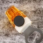 17242105686de7e8443c1ee3292334a062e8086484.jpg - Bling On Decor® | Luxury Handmade Home Decor Close-up of a glass bottle with amber liquid and a large black cap, with a metal bottle opener in the foreground on a textured surface. - Bling On Decor® | Luxury Handmade Home Decor ANJU BALA