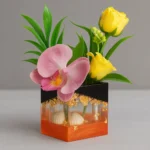 Decorative flower arrangement in a square orange-and-black vase with pink orchid and yellow roses, gold accents, and green leaves. - Bling On Decor® | Luxury Handmade Home Decor ANJU BALA