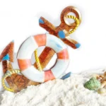 Decorative nautical ornament with an orange crackle-painted anchor and a white-and-pink lifebuoy, tied with a yellow rope on sand. - Bling On Decor® | Luxury Handmade Home Decor ANJU BALA