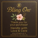 Gold-embossed card with 'Bling On' logo, a pink flower, and the message 'Thank you for your purchase! Handle with Love & Care' on a brown textured background. - Bling On Decor® | Luxury Handmade Home Decor ANJU BALA
