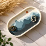 Decorative tray displaying two gold rings and a pearl bracelet on layered blue-toned paper waves, set on a light wooden surface.