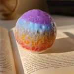 Rainbow-colored bath bomb resting on open book pages, blending pastel hues from purple to red. - Bling On Decor® | Luxury Handmade Home Decor ANJU BALA
