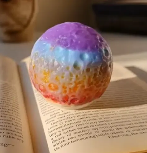 Rainbow-colored bath bomb resting on open book pages, blending pastel hues from purple to red. - Bling On Decor® | Luxury Handmade Home Decor ANJU BALA