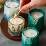 Hand is lighting a decorative botanical candle with a match among blue and green candles on a wooden tray. - Bling On Decor® | Luxury Handmade Home Decor ANJU BALA