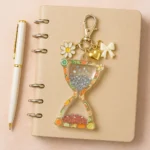 ChatGPT-Image-May-23-2025-08_51_08-PM.webp - Bling On Decorยฎ | Luxury Handmade Home Decor Beige spiral notebook with a decorative charm keychain showing an hourglass filled with glitter, citrus-fruit border, and charms (daisy, heart, bow). Plus a white-and-gold pen nearby. - Bling On Decorยฎ | Luxury Handmade Home Decor ANJU BALA
