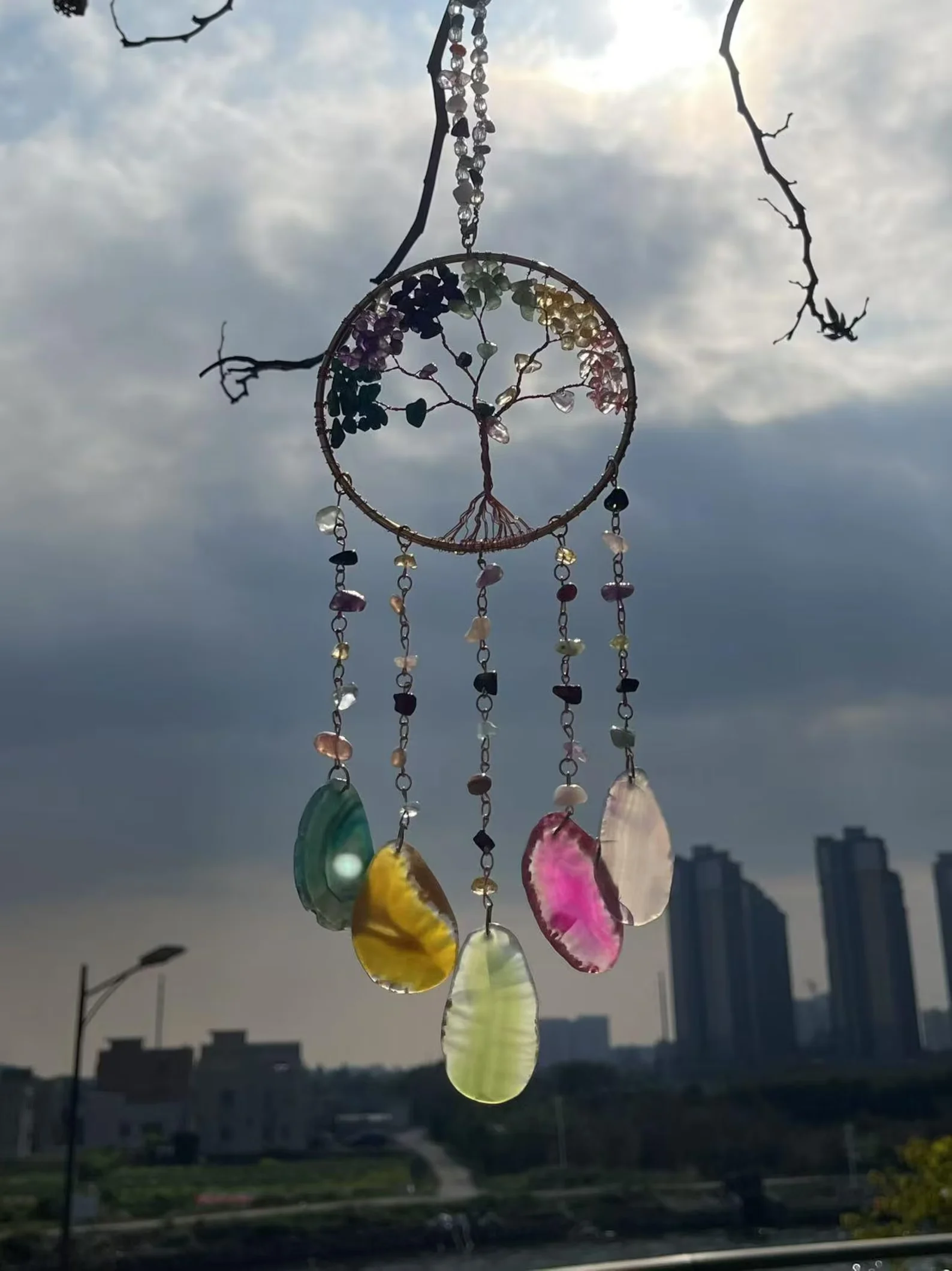 Dreamcatcher hanging in front of a city skyline with colorful bead charms and shells against a cloudy sky. (Informative) - Bling On Decor® | Luxury Handmade Home Decor ANJU BALA