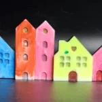 Row of colorful ceramic houses in pastel pink, blue, orange, yellow, and white lined up against a dark background with blue stones in the foreground - Bling On Decor® | Luxury Handmade Home Decor ANJU BALA