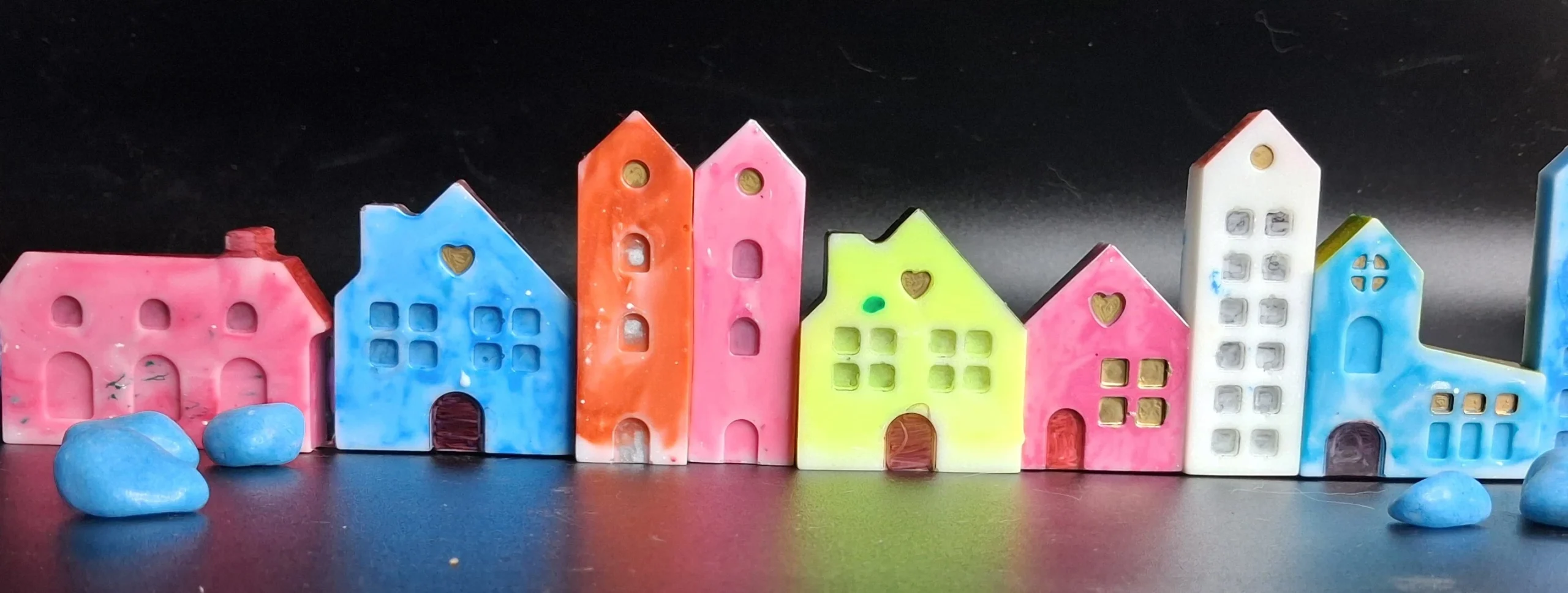 Row of colorful ceramic houses in pastel pink, blue, orange, yellow, and white lined up against a dark background with blue stones in the foreground - Bling On Decor® | Luxury Handmade Home Decor ANJU BALA