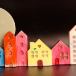 Whimsical row of colorful ceramic houses with a large white crescent moon sculpture behind them on a dark background. - Bling On Decor® | Luxury Handmade Home Decor ANJU BALA