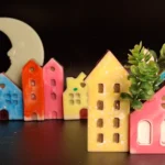 Colorful ceramic town figurines arranged in a row with a large pale crescent moon in the dark background; whimsical decorative scene. - Bling On Decor® | Luxury Handmade Home Decor ANJU BALA