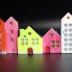 Row of pastel ceramic houses in pink, blue, orange, yellow and white, arranged along a dark surface with blue stones in the foreground. - Bling On Decor® | Luxury Handmade Home Decor ANJU BALA