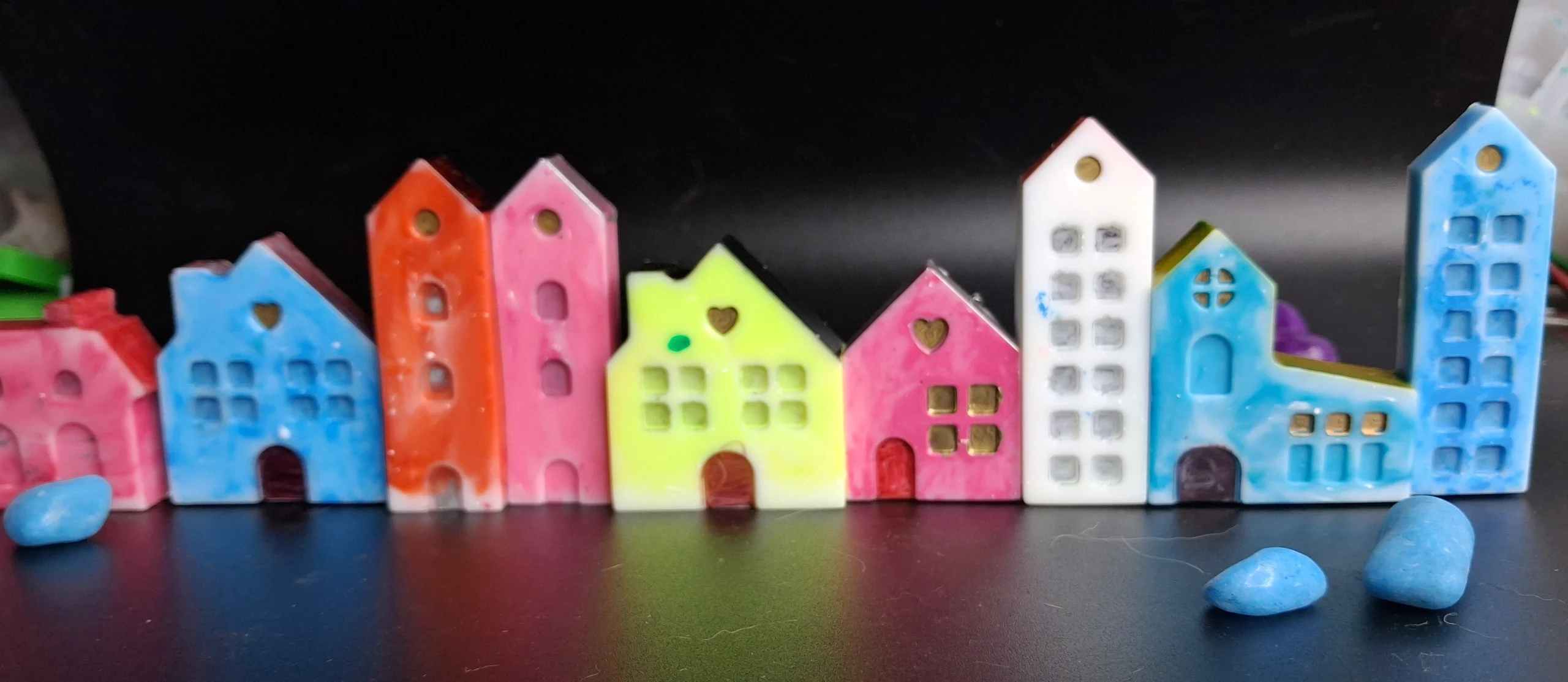 Row of pastel ceramic houses in pink, blue, orange, yellow and white, arranged along a dark surface with blue stones in the foreground. - Bling On Decor® | Luxury Handmade Home Decor ANJU BALA