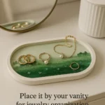 Gold rings and a pearl necklace lie on a white oval vanity tray with a green landscape design. - Bling On Decor® | Luxury Handmade Home Decor ANJU BALA