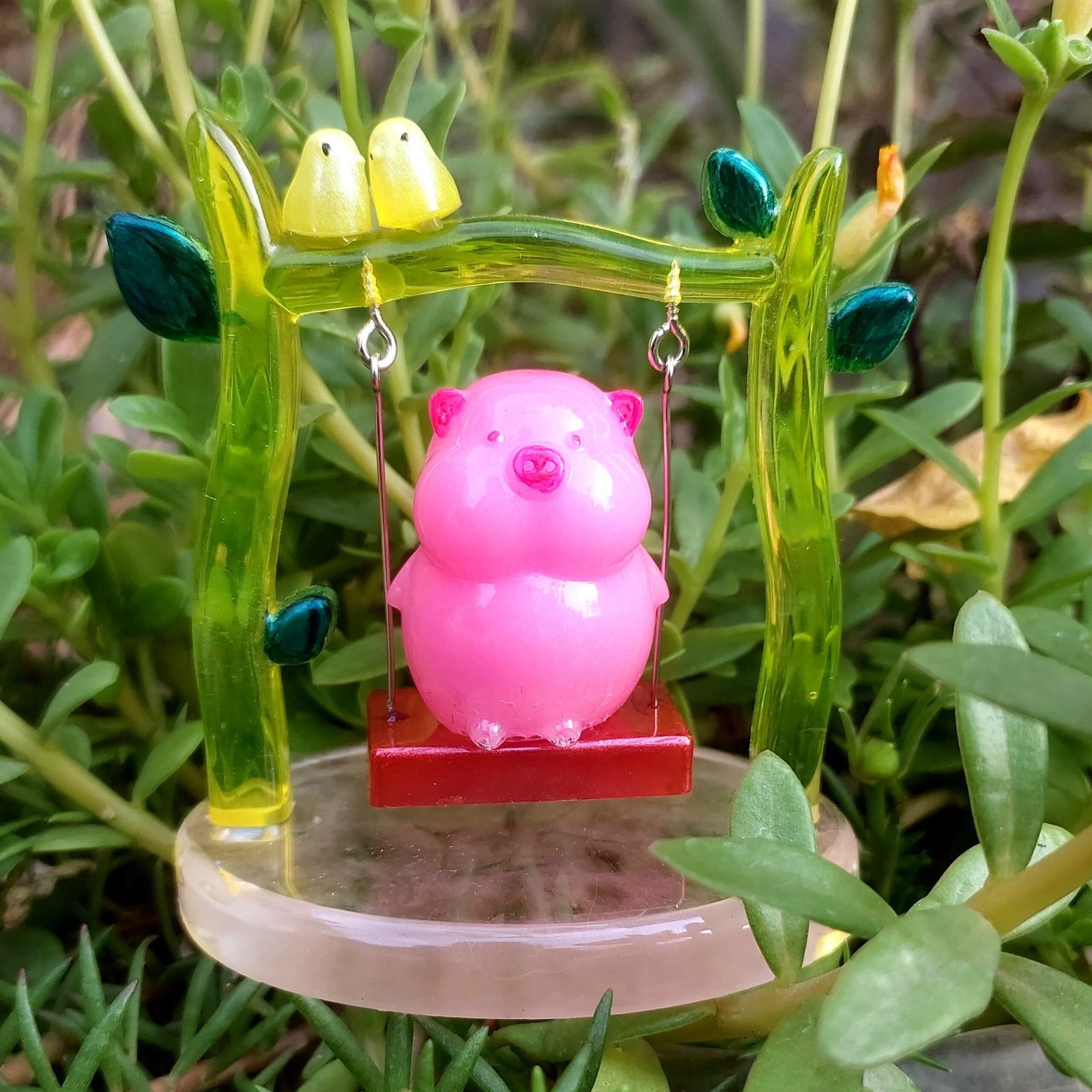 Pink plastic pig figurine hanging on a small red swing, set against a green translucent garden frame and leafy background - Bling On Decor® | Luxury Handmade Home Decor ANJU BALA