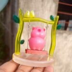 Pink pig figurine hanging inside a translucent yellow-green frame with small metal chains, atop a circular base (ornamental display). - Bling On Decor® | Luxury Handmade Home Decor ANJU BALA