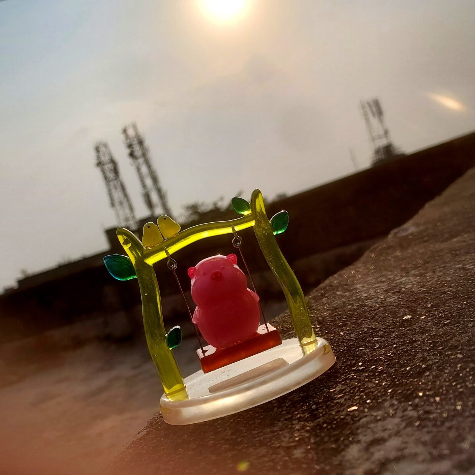 Plastic yellow-green toy crown with a pink pig figure sits on a rough outdoor ledge, sunset sky and distant cell towers in the background. - Bling On Decor® | Luxury Handmade Home Decor ANJU BALA