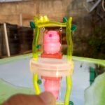 Pink pig toy sitting on a small swing inside a translucent yellow-green frame with green leaf accents, outdoors on a reflective surface. - Bling On Decor® | Luxury Handmade Home Decor ANJU BALA
