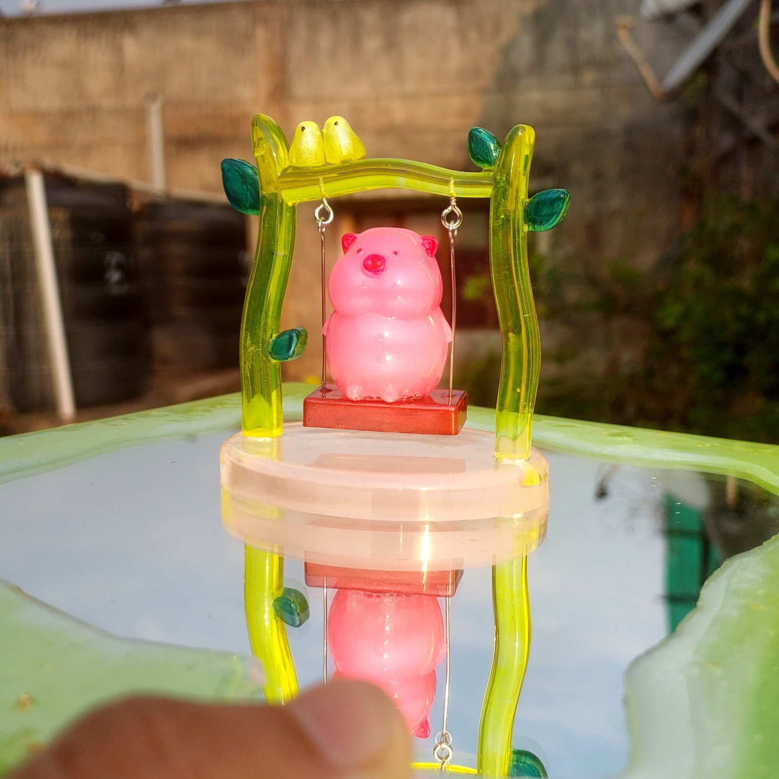 Pink pig toy sitting on a small swing inside a translucent yellow-green frame with green leaf accents, outdoors on a reflective surface. - Bling On Decor® | Luxury Handmade Home Decor ANJU BALA