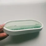 White oval dish held by a hand, containing a translucent green gel-like substance or soap slab embedded inside. - Bling On Decor® | Luxury Handmade Home Decor ANJU BALA