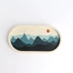 Oval stamp depicting layered blue mountains with a small orange sun in a pale sky. - Bling On Decor® | Luxury Handmade Home Decor ANJU BALA