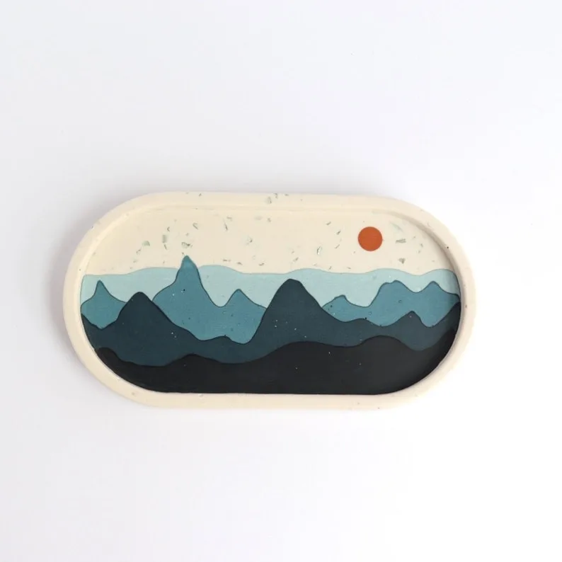 Oval stamp depicting layered blue mountains with a small orange sun in a pale sky. - Bling On Decor® | Luxury Handmade Home Decor ANJU BALA