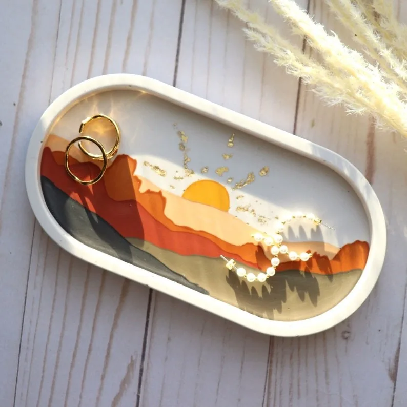 Decorative white oval tray with a desert sunset landscape design, holding two gold rings and a pearl necklace on a light wood surface. - Bling On Decor® | Luxury Handmade Home Decor ANJU BALA
