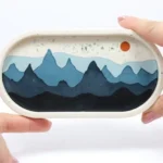 Hand-held oval diorama showing blue layered mountains with an orange sun in a white frame. - Bling On Decor® | Luxury Handmade Home Decor ANJU BALA