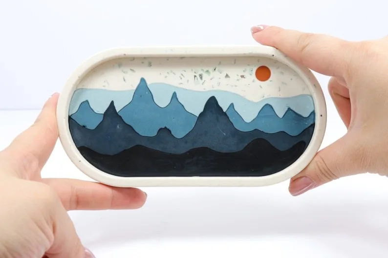 Hand-held oval diorama showing blue layered mountains with an orange sun in a white frame. - Bling On Decor® | Luxury Handmade Home Decor ANJU BALA