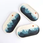 Three oval decorative coasters with a layered blue mountain motif and a small orange dot on a white speckled background - Bling On Decor® | Luxury Handmade Home Decor ANJU BALA