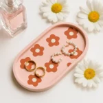 Pink tray with orange flower prints displaying two gold rings and pearl hoop earrings, with a perfume bottle and white daisies nearby.