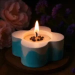 Flower-shaped candle with a single lit wick, blue and white gradient, resting on a wooden tray with blurred flowers in the background - Bling On Decor® | Luxury Handmade Home Decor ANJU BALA
