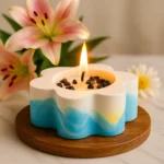 Flower-shaped candle with a lit wick on a wooden round tray, blue and white gradient, with pink lilies in the background. - Bling On Decor® | Luxury Handmade Home Decor ANJU BALA