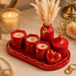 Red decorative candle centerpiece: four red hexagonal candles on a red tray with a heart ornament, red ribbed jar vase with dried pampas, pink rose, and gold jewelry nearby - Bling On Decor® | Luxury Handmade Home Decor ANJU BALA