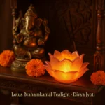 Glowing orange lotus-shaped tealight on a wooden altar with a brass Ganesha statue in the background and orange marigold flowers nearby, creating a warm devotional scene. The image includes the text 'Lotus Brahamkamal Tealight - Divya Jyoti' on the bottom. - Bling On Decor® | Luxury Handmade Home Decor ANJU BALA