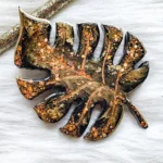Dark, leaf-shaped resin pendant with orange glitter specks, resting on a pale surface with a small branch nearby. - Bling On Decor® | Luxury Handmade Home Decor ANJU BALA