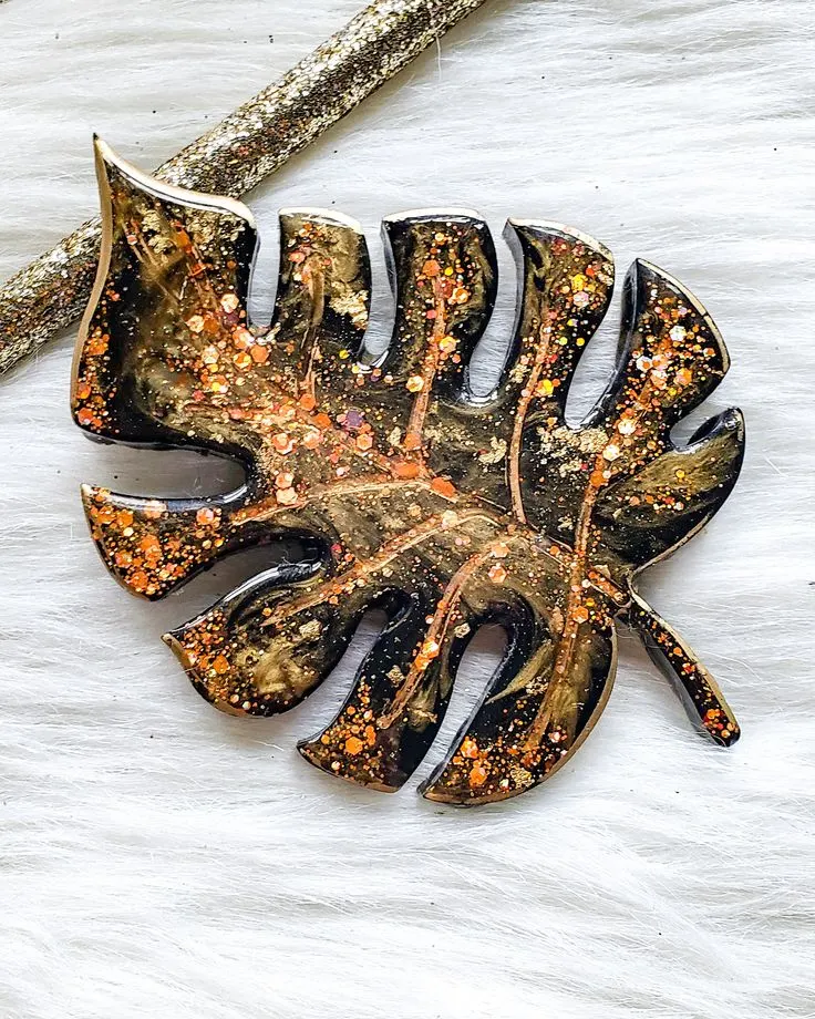 Dark, leaf-shaped resin pendant with orange glitter specks, resting on a pale surface with a small branch nearby. - Bling On Decor® | Luxury Handmade Home Decor ANJU BALA