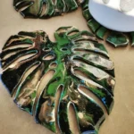 Glossy green and black monstera leaf-shaped decorative piece on a beige surface with cut-out sections and powdery white specks. - Bling On Decor® | Luxury Handmade Home Decor ANJU BALA