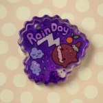 Purple glitter keychain with 'Rain Day' text and a cute brown character beside pink frosting-like details, against a light polka-dot background. - Bling On Decor® | Luxury Handmade Home Decor ANJU BALA