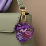 Purple scalloped acrylic tag hanging from a light green handbag; it says 'Rain Day' with a cute smiling berry character and small clouds/leaves in purple tones. - Bling On Decor® | Luxury Handmade Home Decor ANJU BALA
