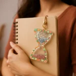 Resin-Handmade-Vintage-Sand-Time-Glitter-Epoxy-Fruit-Hourglass-Keychain-diary-journal-for-time-manment-.webp - Bling On Decorยฎ | Luxury Handmade Home Decor Person holding a beige spiral notebook with a decorative hourglass charm filled with blue stars and red glitter visible on the front cover accessory side - Bling On Decorยฎ | Luxury Handmade Home Decor ANJU BALA