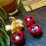 Resin-Love-Cup-table-Top-Desktop-Item-5.webp - Bling On Decor® | Luxury Handmade Home Decor Close-up of red circular containers holding glass marbles on a dark wooden surface, with seashells and plant leaves nearby. - Bling On Decor® | Luxury Handmade Home Decor ANJU BALA
