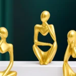 Three gold abstract humanoid sculptures seated and posing on a white surface against a dark teal background. - Bling On Decor® | Luxury Handmade Home Decor ANJU BALA