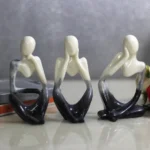 Three abstract ceramic figures in a row, hands cupping their faces, fading from cream to deep blue, on a reflective surface with pink roses and books nearby. - Bling On Decor® | Luxury Handmade Home Decor ANJU BALA