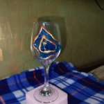 Unleash-the-Sparkle-Blue-Geode-Wine-Glass-4.webp - Bling On Decor® | Luxury Handmade Home Decor Decorated wine glass with blue and copper abstract artwork, resting on a pale pink napkin on a blue patterned surface. - Bling On Decor® | Luxury Handmade Home Decor ANJU BALA
