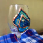 Unleash-the-Sparkle-Blue-Geode-Wine-Glass-9.webp - Bling On Decor® | Luxury Handmade Home Decor Close-up of a colorful hand-painted glass ornament with blue, teal, and copper accents inside a clear glass container. - Bling On Decor® | Luxury Handmade Home Decor ANJU BALA