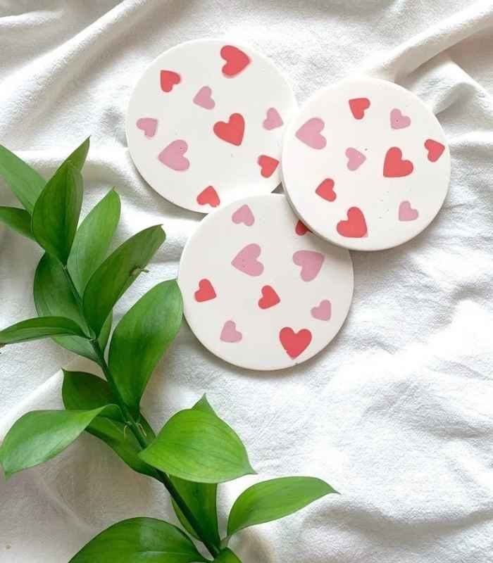 Three white circular cards with red and pink hearts laid on white fabric next to a green leafy branch, conveying a Valentine's theme. - Bling On Decor® | Luxury Handmade Home Decor ANJU BALA
