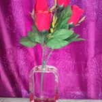 Bouquet of red roses with green leaves in a clear glass vase on a table, against a purple patterned backdrop. - Bling On Decor® | Luxury Handmade Home Decor ANJU BALA