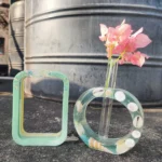 Pink bougainvillea flowers in a tall glass vase placed on the asphalt beside a green, shell-decorated circular sculpture, with a large metal tank in the background. - Bling On Decor® | Luxury Handmade Home Decor ANJU BALA